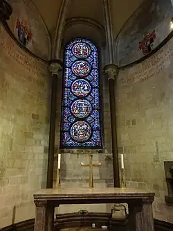 A semi-circular area with stone walls, a domed ceiling and a tall stained glass window with prominent circular decorations.