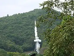 Rocket with smoke in front of green mountain.