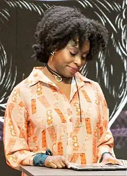 Woman standing at a podium. There is a tablet on the podium from which she is reading.