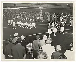 Black and white photo of spectators watching girls march on the street. A group of girls at the front hold two American flags. Girls march in rows further behind. All wear white caps and dark buttoned tops, with white skirts, socks, and shoes.
