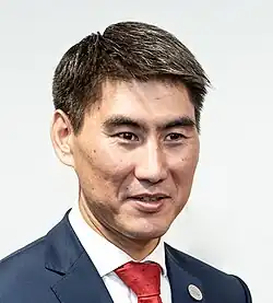 Photograph of Aidarbekov on a light background. He is wearing a dark blue suit jacket with a red tie, and his hair is cut short.