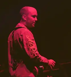 A man with a skinhead haircut sat in front of a musical keyboard. He is looking to his right, away from the camera and is shown side-on. He is wearing a jacket with embroidering on the right sleeve and is holding a drinks can in his right hand. He is bathed in red stage lights.