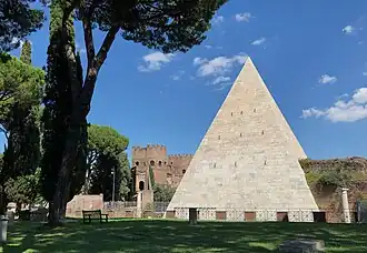 Stone pyramid standing in grassy area with tall trees and red brick fortress in background under blue sky