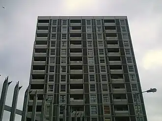 Eleven storeys of a gray residential tower, with a spiked fence and a lamp in the foreground.
