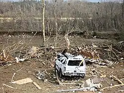 A damaged car sits in front of a row of damaged trees and debris.