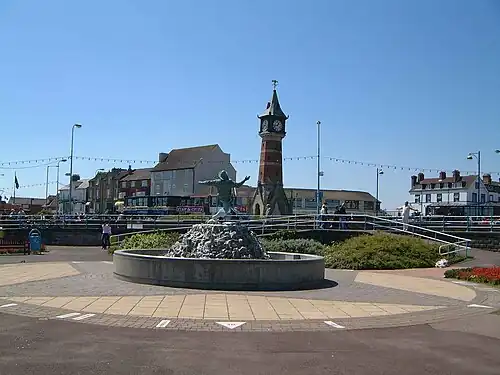 A fountain with a sculpture of a person; a clock tower, people and buildings are in the background