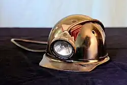 Protective miner's hard hat with an attached electric headlamp, resting on a dark surface.