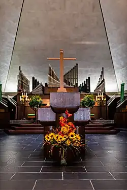 The west wall of the sanctuary, with pulpit and organ. There are flowers in front of the pulpit, a wooden structure with a cross on it. The floor is tiled. Behind the pulpit there is a set of steps ascending to the organ, which has a series of pipes of varying height.