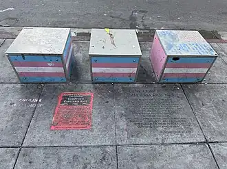 Two plaques embedded into the sidewalk in front of three cubes painted as the trans pride flag.