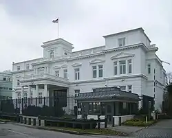 A multi-storey, stand alone white building with a high fence surrounding it. The entrance has a porch with 4 columns.