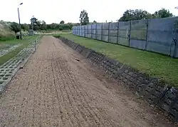 View along a ploughed bare earth strip, the right-hand side of which descends into a ditch faced with concrete. A metal fence is situated further to the right.