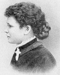 black and white photograph of a white woman with dark, medium length hair, in profile, wearing a dark dress with a white cross on her lapel.