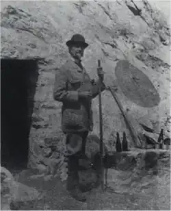 Black and white photo, a man stands holding a walking stick.