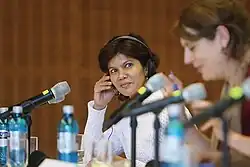 Shreen Abdul Saroor is pictured seated at a panel discussion in 2014. She is seen looking at the woman in the right of the image. Saroor is wearing a white sweater and a headset, which she is holding to her ear with her right hand. In the foreground of the picture are water bottles and microphones.