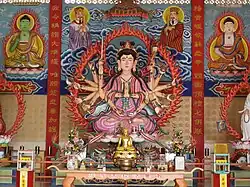 Statue of Zhunti Guanyin with eighteen arms wielding different dharma instruments or displaying mudras, flanked by devas and with Yaoshi Fo (Bhaiṣajyaguru) on her left and Amituofo (Amitābha) on her right. The Chinese text on the front of the altar reads, Namo Zhunti Wang Pusa, or "Namo Cundi King Bodhisattva." Enshrined at a Chinese Buddhist temple in Kelantan, Malaysia.