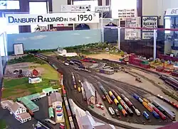 A model train display with the station building at the left lower corner with various types of trains on the tracks next to the station and the railyard. A sign above it, on the upper left, reads "Danbury Railyard in the 1950s"