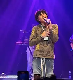 DannyLux performing at Syracuse University on October 19, 2024. He is wearing an army-green shirt, a chain with diamond encrusted castle, blue pants, and diamond earrings