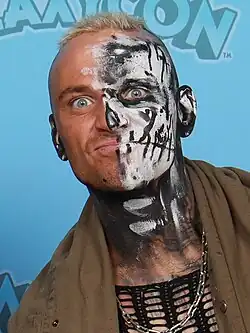 A blonde man wearing face paint and a brown jacket.