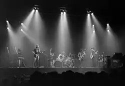 A monochrome image of members of the band. The photograph is taken from a distance, and is bisected horizontally by the forward edge of the stage. Each band member and his equipment is illuminated from above by bright spotlights, also visible. A long-haired man holds a guitar and sings into a microphone on the left of the image. Central, another man is seated behind a large drumkit. Two men on the right of the image hold a saxophone or a bass guitar and appear to be looking in each other's general direction. In the foreground, silhouetted, are the heads of the audience.