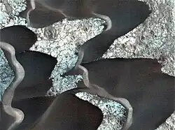 Dark sand dunes on Nili Patera of probable volcanic origin. The lighter-coloured surface is ancient lava, which has cooled. The cracks on the lava floor are filled with dark, volcanic sand.