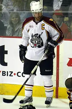 A Caucasian ice hockey player standing on the ice looking down. He wears a white helmet, white jersey with maroon shoulder and arm stripes, black shorts, and white socks. The jersey has a stylized wolf head with a stick and puck behind it as the logo.