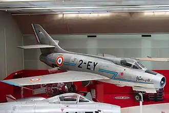 A color photograph of a metallic silver-colored jet numbered 2-EY in a museum. Another similar aircraft can be seen on the bottom.