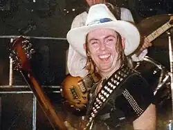 A man with shoulder-length hair wearing a black T-shirt & a white cowboy hat, with a studded guitar strap