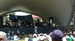 Performers on an outdoor stage with shell-shaped overhang before an audience seated in the sun.