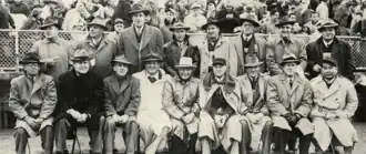 Black-and-white Ben Day image with two rows of well-dressed men in trench coats and trilbies; eight men are standing in the back, nine are sitting in front of them; behind them all is a fenced-in crowd