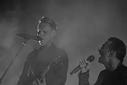 A black and white photograph showing a man holding a guitar on the left, and another man singing into a microphone on the right, with a haze covering the whole photograph.