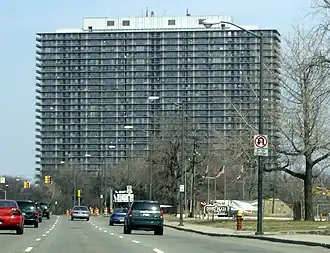 A low and wide skyscraper building of between twenty and thirty stories in height. In the foreground is a road lined with deciduous trees.