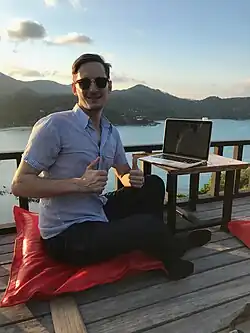 A man with a laptop sitting on a wooden balcony overlooking a tropical bay and green mountains.