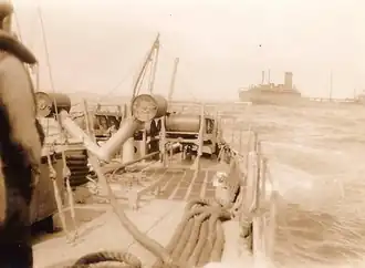 An oil tanker is half-visible just behind the stern of the vessel; the tanker is partially cut out by the camera's frame. On the ship is a sailor, and behind him a Y-gun used to launch depth charges.