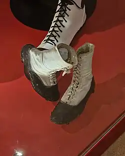 a pair of worn clown shoes on a red plinth. The shoes feature shiny black leather rim around the sides of the foot, with the upper part of the shoe white fabric with white laces