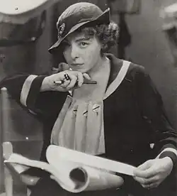 Woman in cloche-style hat and dark coat holds a pen and rolled papers while looking intently forward