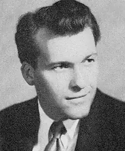 A monochrome bust photo of a white man with dark hair, he is wearing a dark-colored suit and tie, looking to the camera's right.