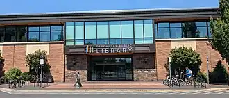 Photograph of the Downtown Bend Library, a modern brick and glass building, on a sunny day. Above the doors is a sign that reads Deschutes Public Library and a minimalist logo of multicolored vertical lines representing books. A metal statue appears in front of the library. There are bike racks on either side of the entrance. Two people approach the library afoot.