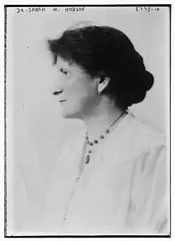 A white woman of middle age, with dark hair dressed to the nape, photographed in profile wearing a white blouse and a beaded necklace