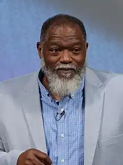Headshot of a bearded Baucham in a shirt and jacket, standing in front of a gray background