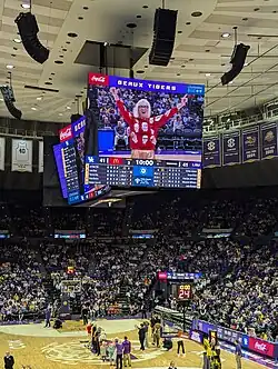 In a basketball stadium, the jumbotron shows a person smiling in a blonde wig and red patterned top. A number of colorfully dressed people are lined up on the basketball court.