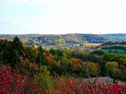 Landscape with low rolling hills, farm buildings in the middle distance surrounded by fields, and bright early autumn foliage