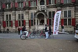 People arriving at and leaving Delft City Hall on election day 2025. In front of the historic building is a portico with stairs on both sides and a temporary accessibility ramp fitted over the right staircase. A row of parked bicycles stands in front of the portico, with two cyclists just arriving.