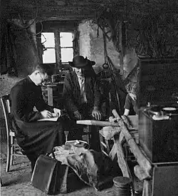 Three white people seated in a barn in 1939; a man in priest's clothing, a farmer at center, and a woman with her head down, writing; a recording device is propped on an overturned wheelbarrow in front of the group.