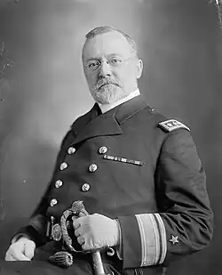 An elderly man with a bushy beard and glasses looking at the camera. He is wearing a military uniform and clutching a sword.