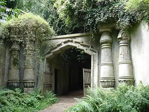Weathered stone entrance with Egyptian Revival columns overgrown with ivy and ferns leading into shadowy passageway