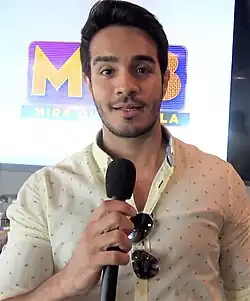 close-up of Ektor Rivera wearing a light-colored patterned shirt with aviator sunglasses hanging from the neck, holding a microphone, and looking directly at camera with mouth slightly open