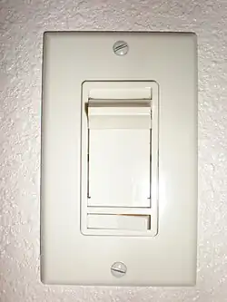 Vertical rectangular plastic cover plate with vertical slider and a smaller horizontal rocker switch below. Two exposed slotted screw heads hold the cover plate to the wall box.