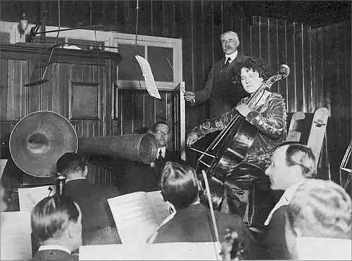 A black and white image of a cellist recording with an orchestra, with the composer conducting