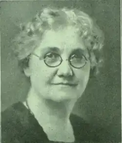 1930 B&W portrait photo of an old woman with wavy white hair and glasses, wearing a dark blouse.