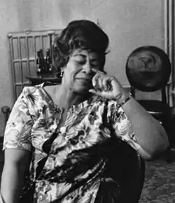 Fitzgerald is sitting down on a chair inside a home. She is wearing a casual dress and leaning over.